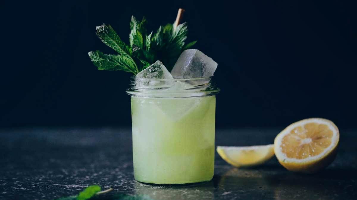 This is a glas filled with a green and yellow cocktail, decorated with ice cubes, lemon slices and lemon balm leaves